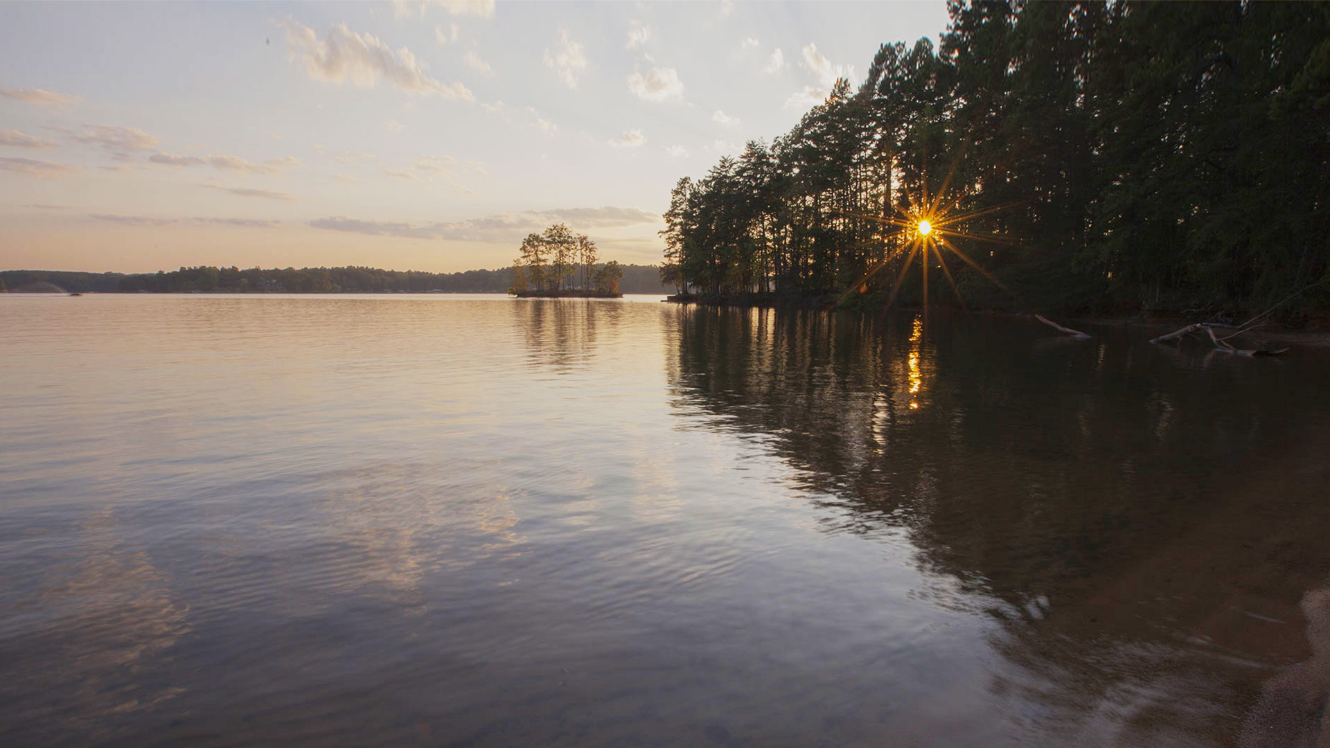 A tranquil lakeside scene at dusk, with a clear reflection of the sun in the water and a calm shoreline.