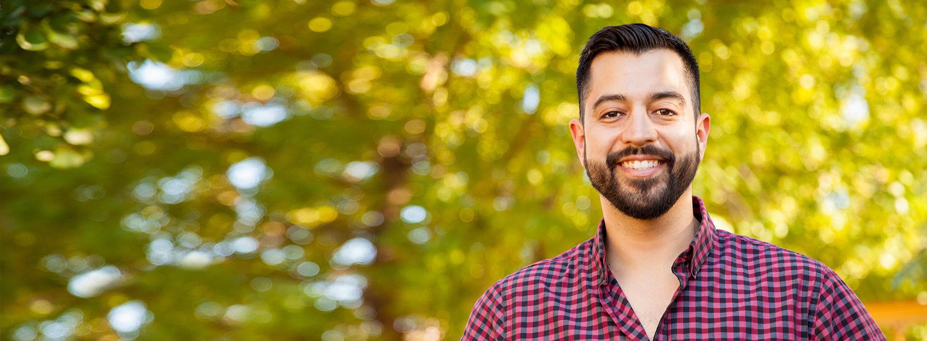 The image shows a man standing outdoors, smiling at the camera. He is wearing a checkered shirt and appears to be in a park-like setting with trees behind him.
