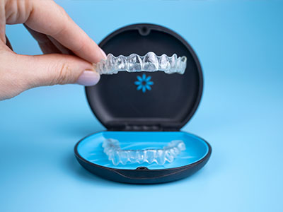 A person s hand is holding a clear plastic tray with a set of white dental implants, which are placed inside a blue and black box labeled DENTAL IMPLANTS on a light blue background.